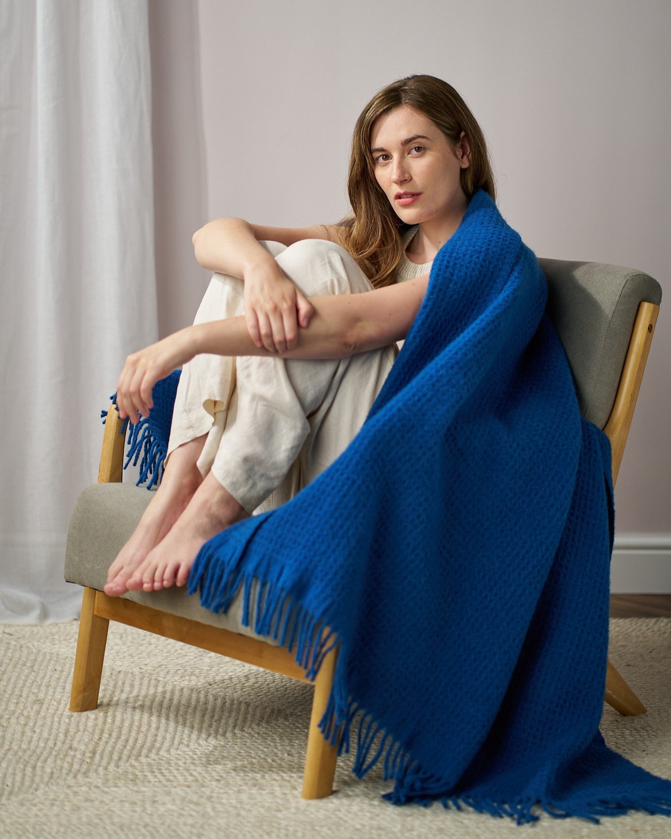 Young woman on a wooden chair wrapped in the Totterdown Blanket Gift Box: Curious, showcasing its chunky texture and fringed edges in a cozy setting.