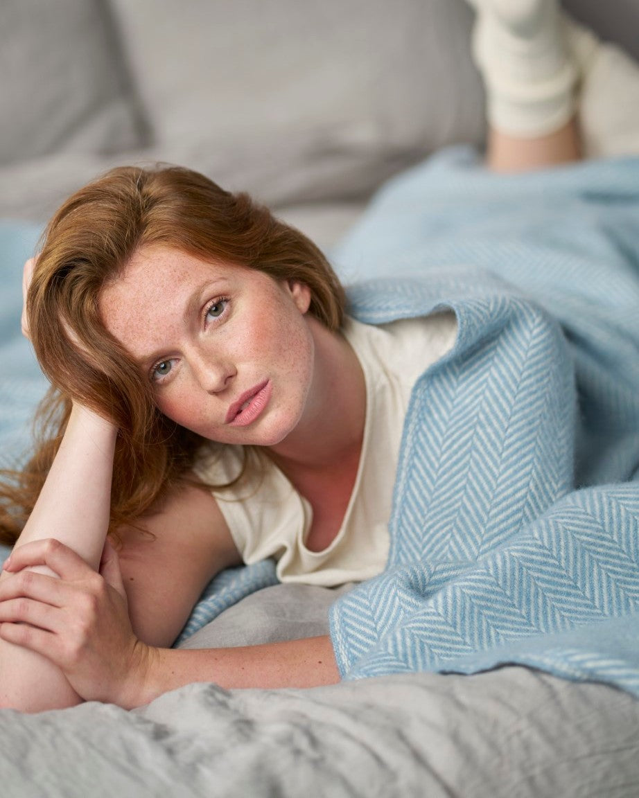Young woman on bed under Clifton Blanket Hamper: Harbourside, showcasing its woven herringbone pattern, creating a relaxed, intimate atmosphere.