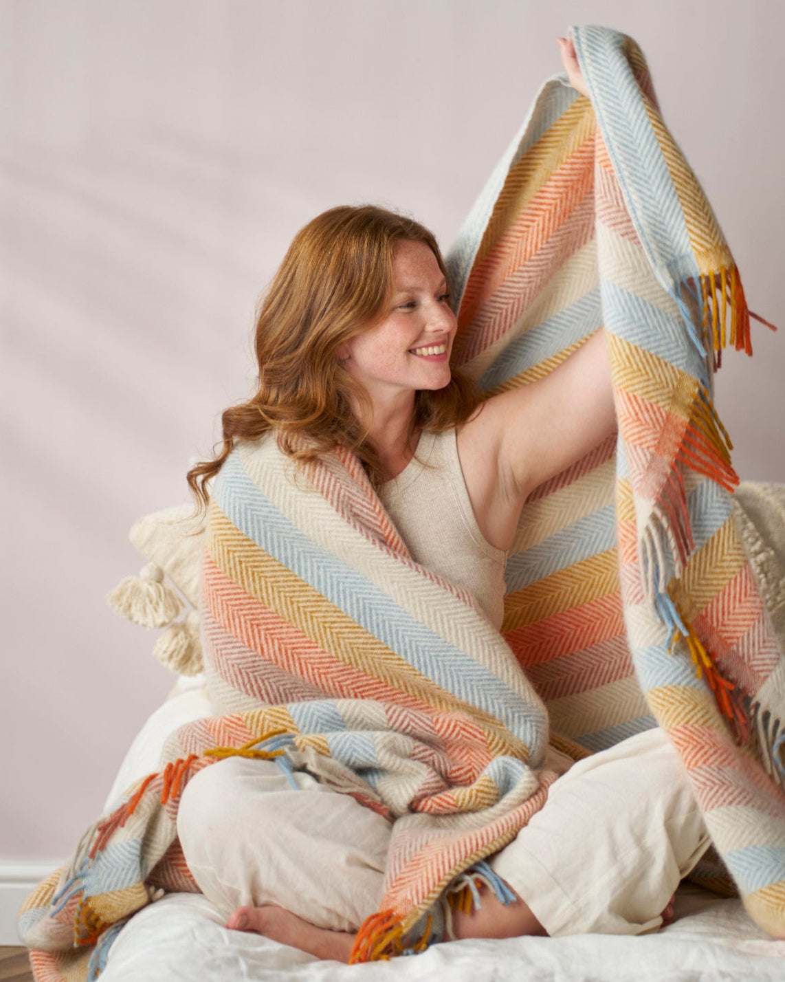 Young woman wrapped in a Rainbow Stripe Blanket from the Boho Gift Box, showcasing its herringbone pattern and fringed edges in a cozy setting.