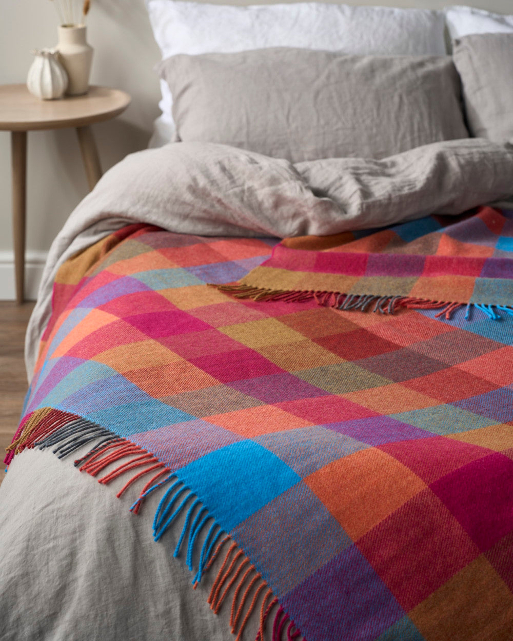 Merino Check Blanket: Multicolour draped over a bed, showcasing its vibrant check pattern and soft texture. Ideal for adding a joyful touch to any space.
