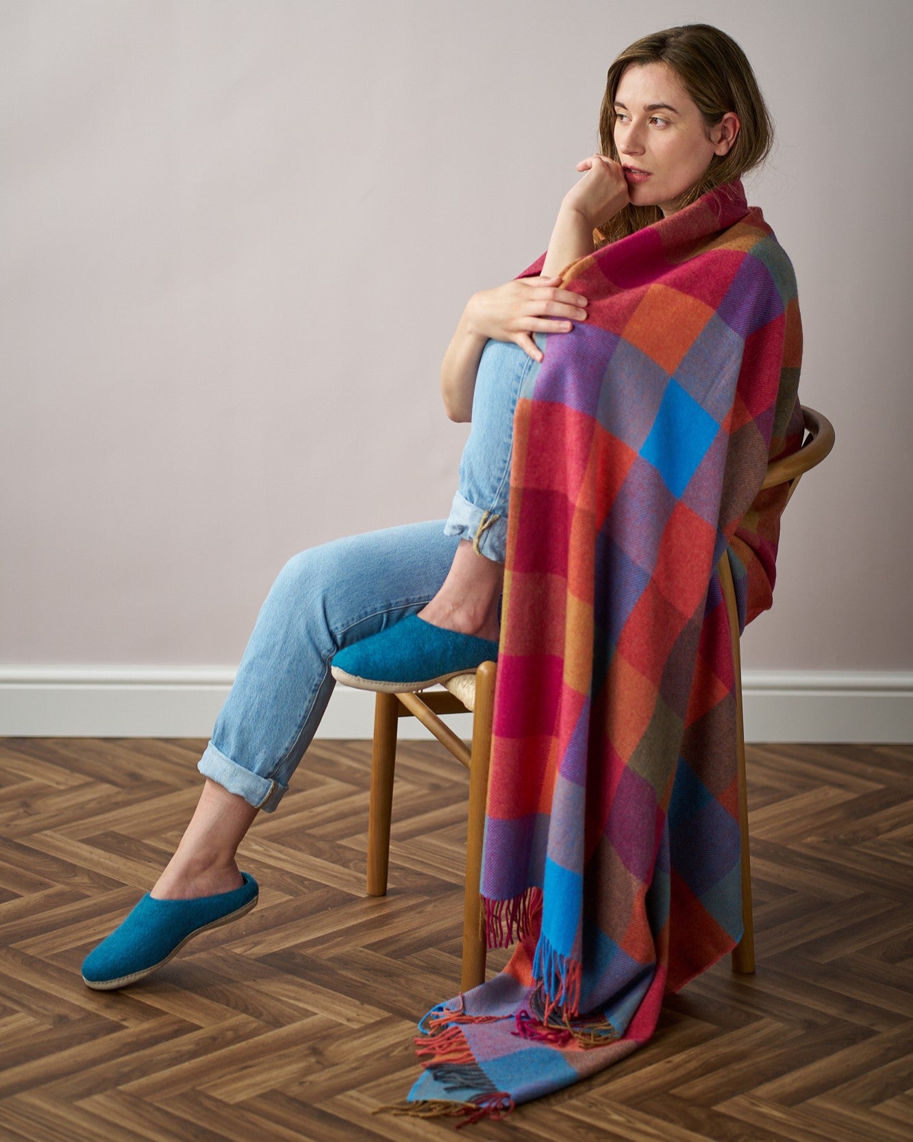 Woman seated with a Merino Check Blanket: Multicolour, draped over her, showcasing its vibrant, soft texture.