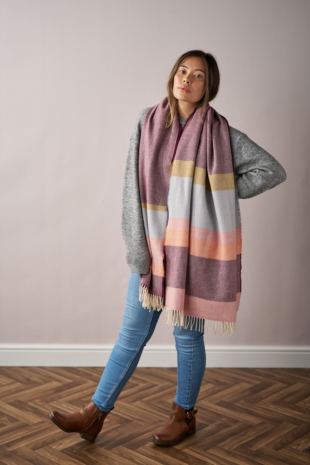 Woman wearing the Oversized Blanket Scarf: Berry Stripe, made of merino lambswool, standing against a wall, showcasing its cozy wrap style.