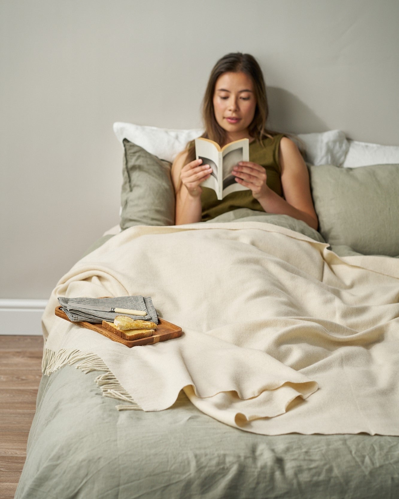 Person reading in bed with a Merino Herringbone Blanket: Cream draped over sage-green sheets, enhancing the cozy, elegant setting with its soft, luxurious texture.