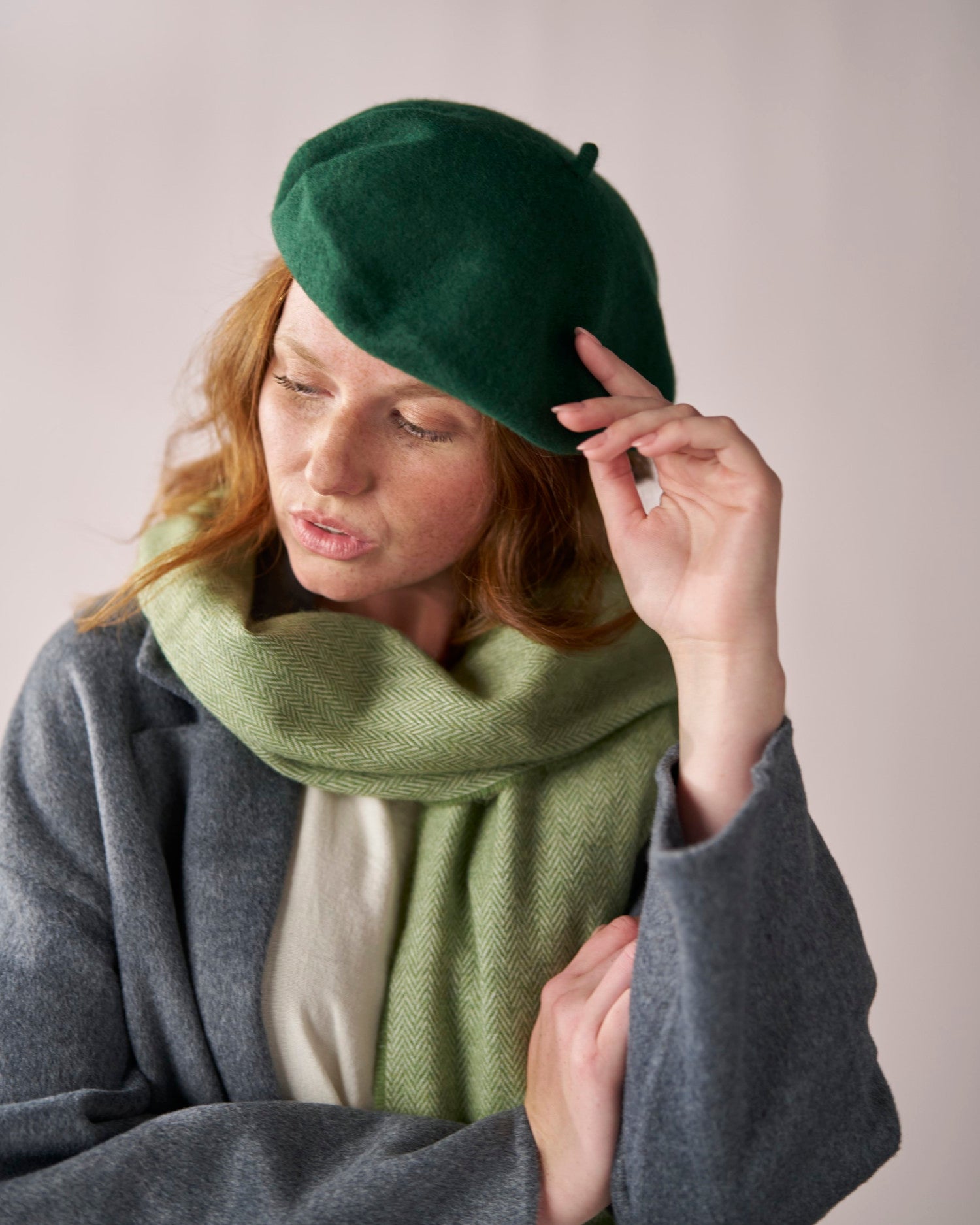 Young woman wearing the Wool Beret Hat: Dark Green, showcasing its thick, premium wool texture. She touches the beret lightly, paired with a herringbone scarf.