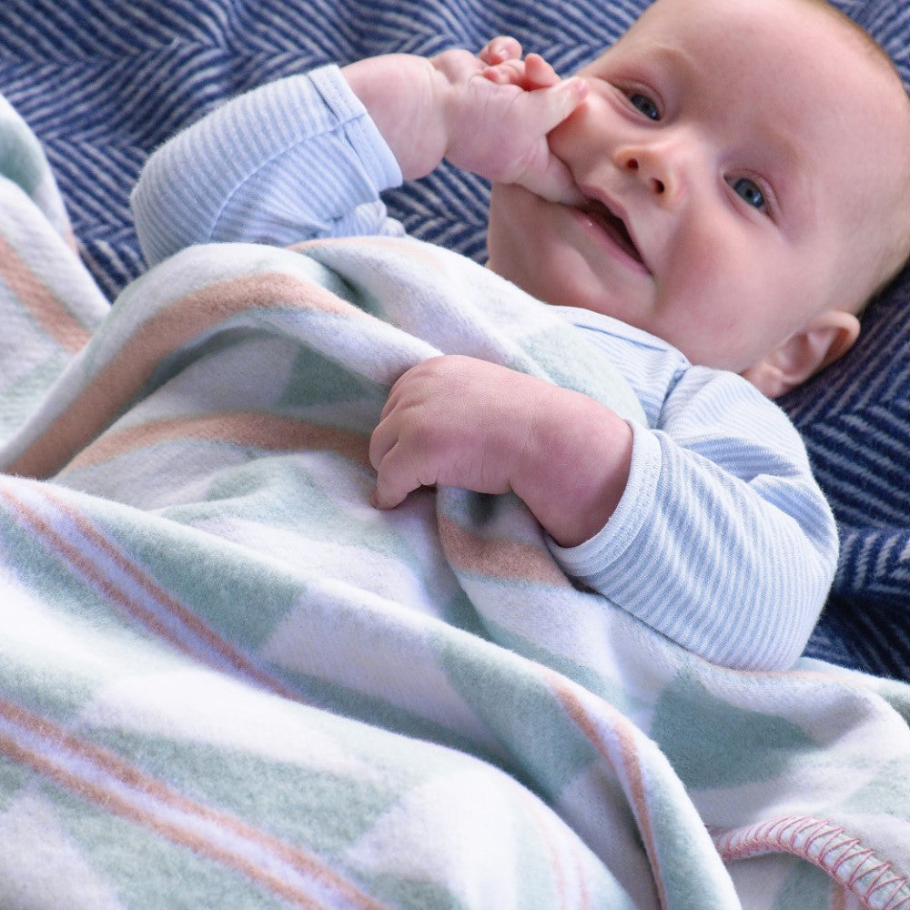 Baby lying on a Cloud Cotton Baby Blanket: Harmony Celadon, featuring a geometric design with a contrasting blanket-stitched border, perfect for sensitive skin.