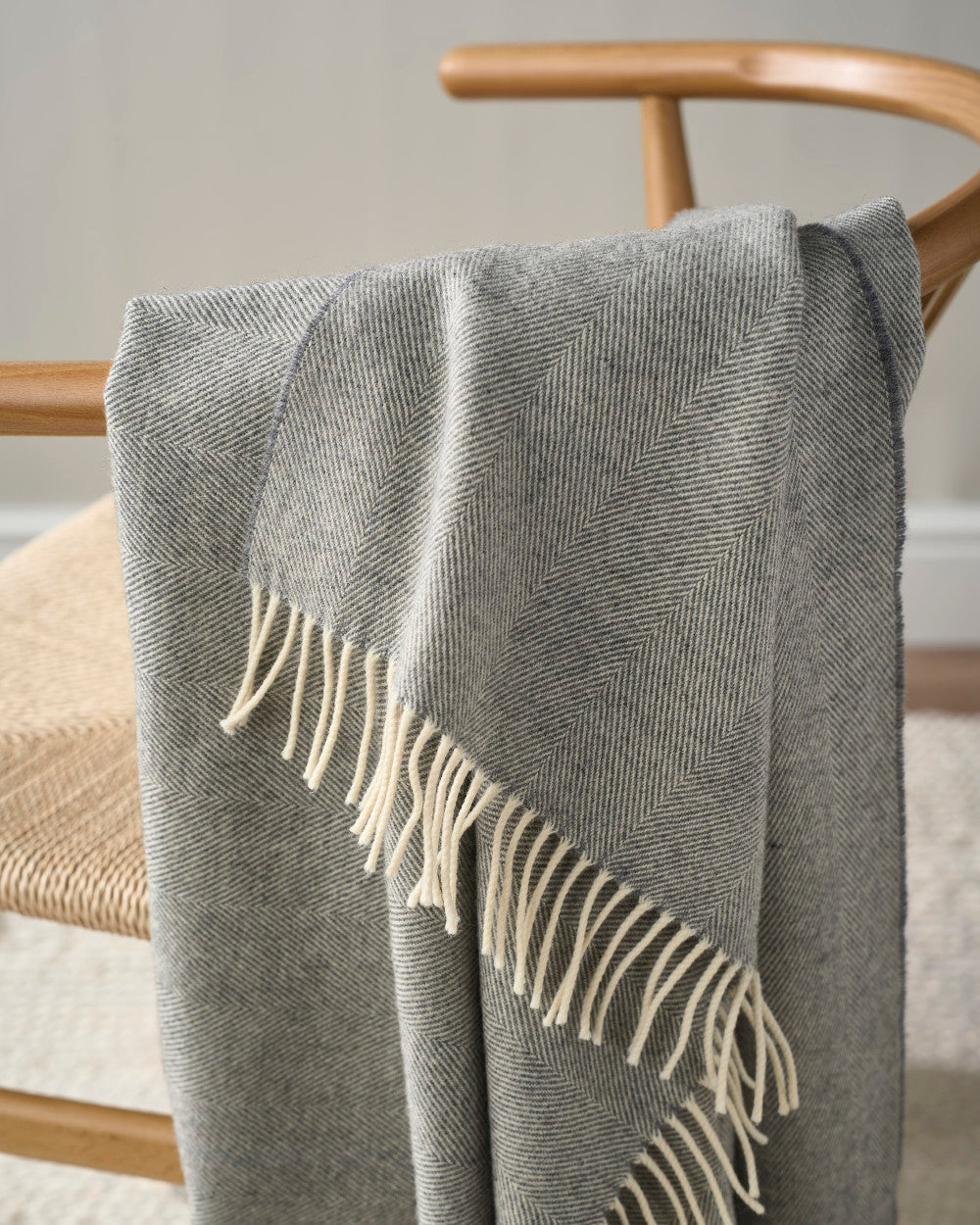Merino Blanket Gift Box: Uniform Grey, featuring a light-gray herringbone-patterned throw with fringe, draped over a wooden chair in a cozy setting.