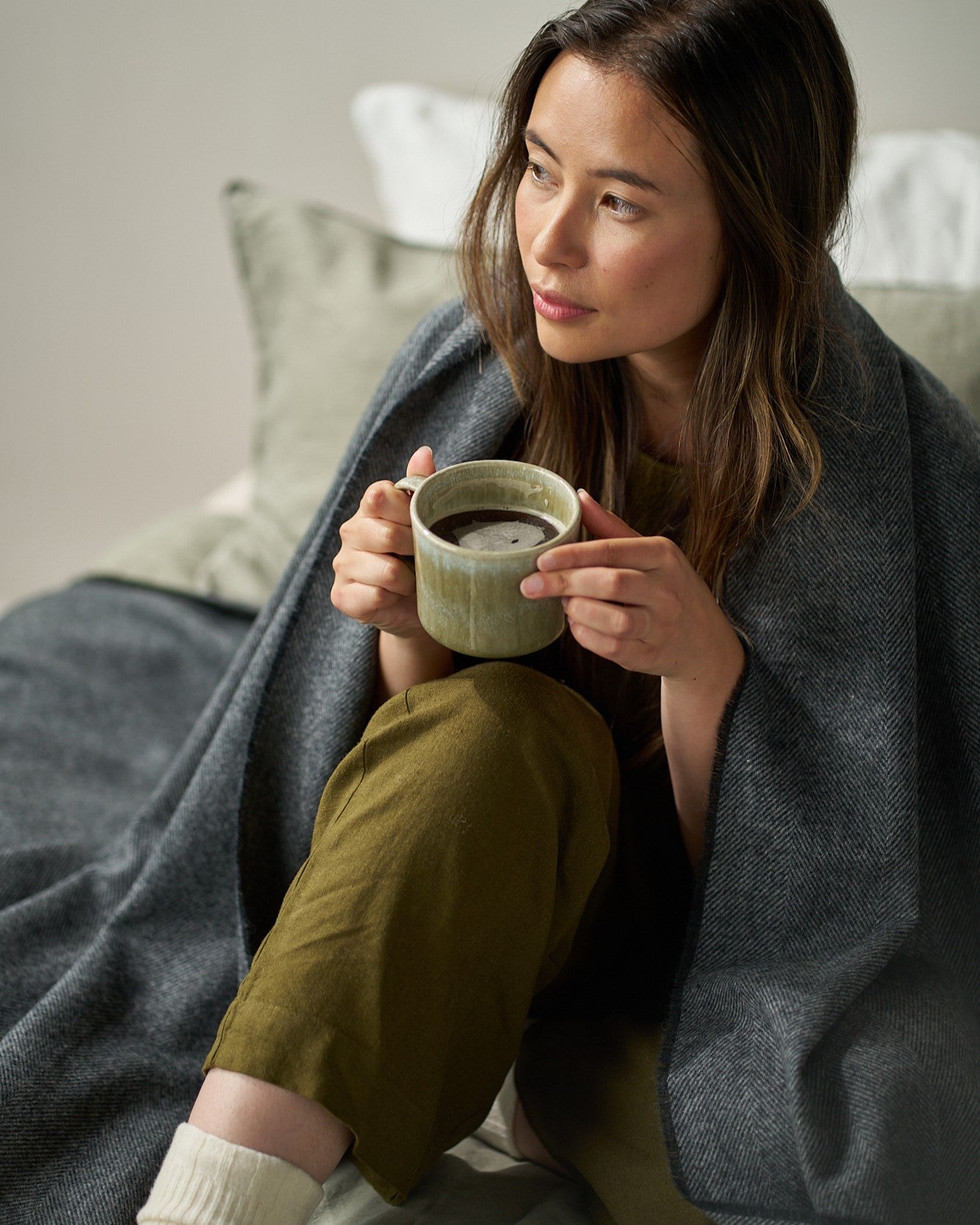 Person wrapped in a Graphite Grey Merino Blanket, holding a mug. Part of the Merino Blanket Gift Box, featuring comfort and relaxation essentials.