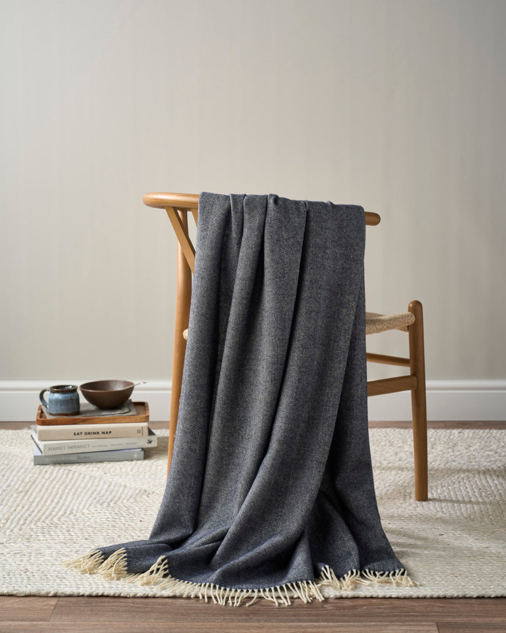 Merino Blanket Gift Box: Navy Blue displayed on a mid-century chair, featuring a woven seat, alongside a cozy setup with books, a ceramic mug, and a bowl.