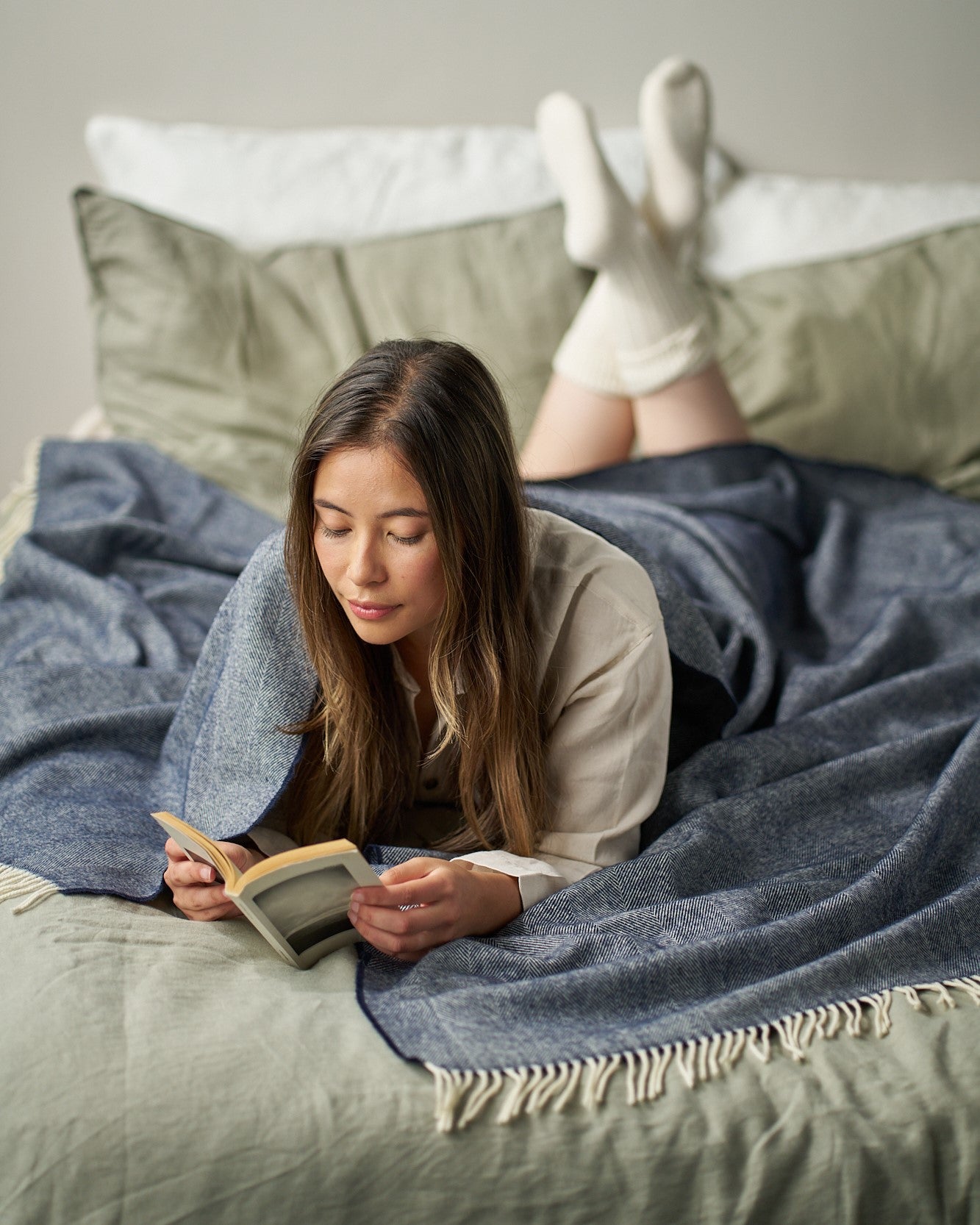 Woman reading on bed with a dark-blue Merino Blanket Gift Box: Navy Blue covering her, surrounded by cozy pillows, creating a serene atmosphere.