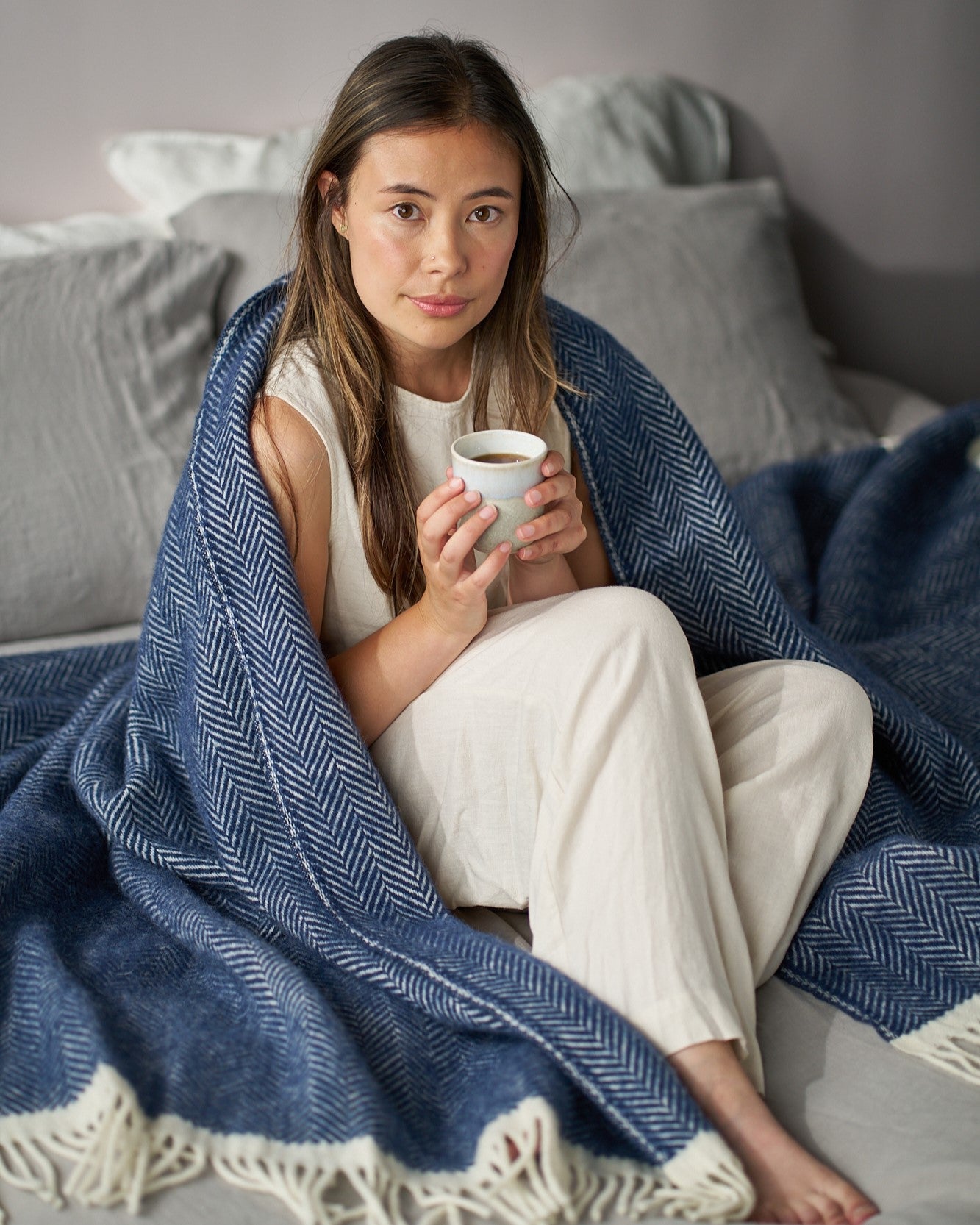 Young woman wrapped in the Clifton Herringbone Blanket: Brunel, holding a mug, sitting among neutral pillows, portraying a cozy, relaxed scene.