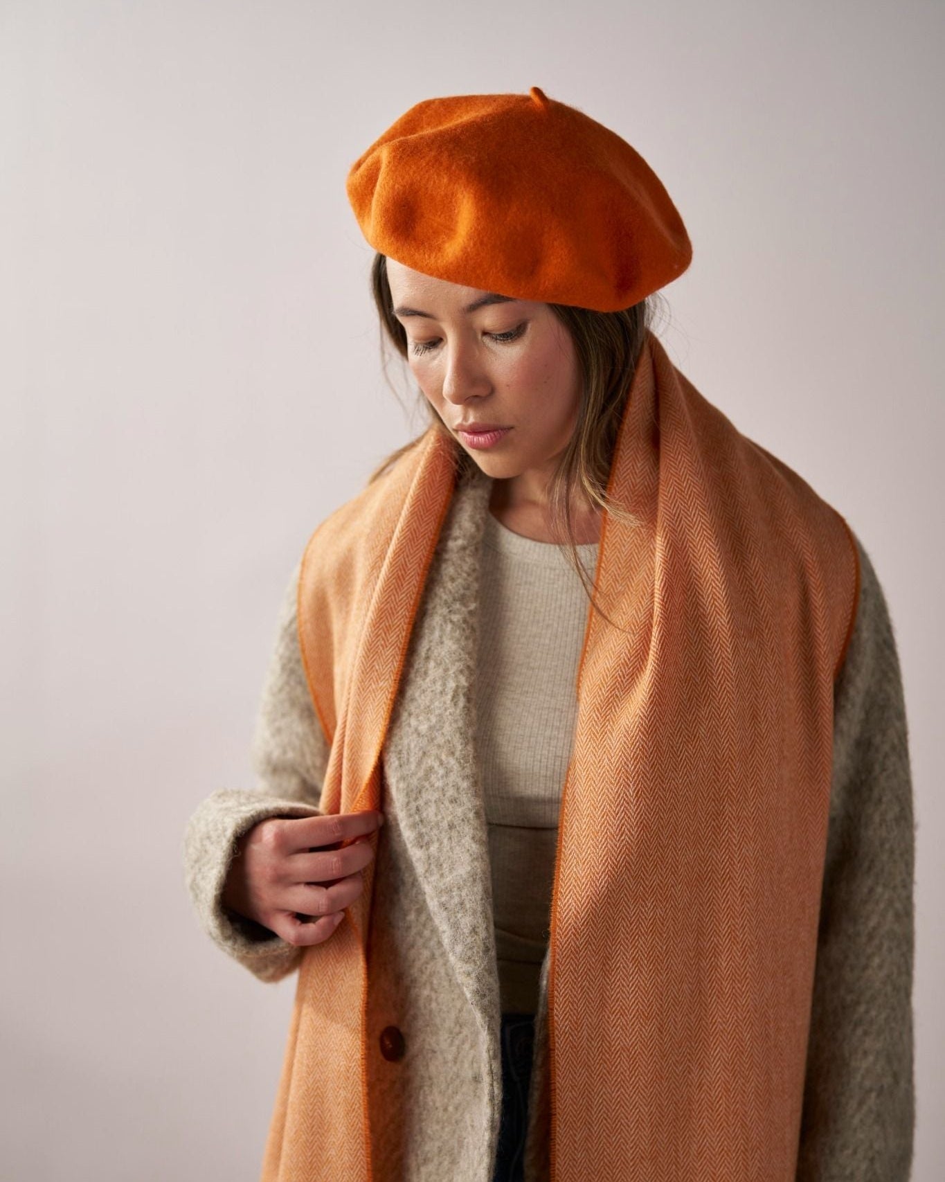 Model wearing the Wool Beret Hat: Orange, a stylish premium felt hat, paired with a matching scarf and gray wool coat in an autumnal setting.