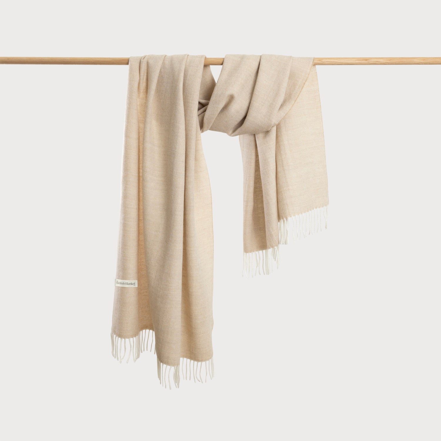 Oversized Blanket Scarf Gift Box: Basswood Herringbone, featuring a light-beige, fringed merino wool throw draped over a wooden rod, showcasing its herringbone weave.