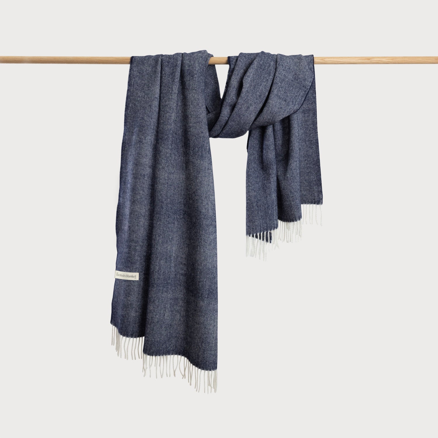 Oversized Blanket Scarf: Navy Blue wool wrap with white fringe, draped over a wooden rod, styled as a cozy throw or shawl.