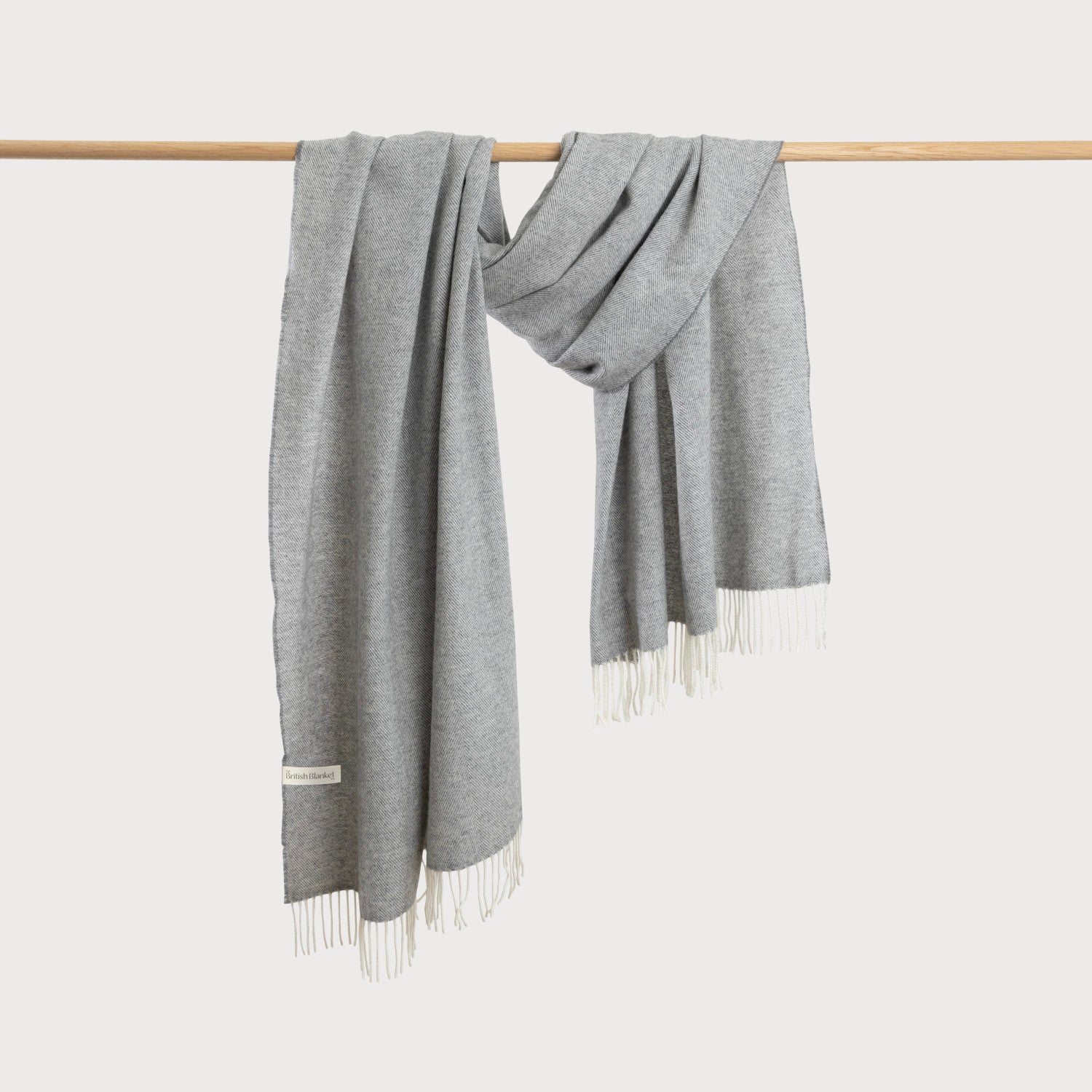 Oversized Blanket Scarf Gift Box: Uniform Grey, featuring a long, light-gray woven wrap with herringbone texture, white fringe, and a woven label, draped over a wooden rod.
