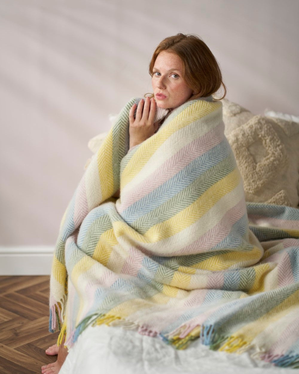 Young woman wrapped in a Rainbow Stripe Blanket Gift Box: Pastel, sitting relaxed with toes on wood floor, against a neutral backdrop.