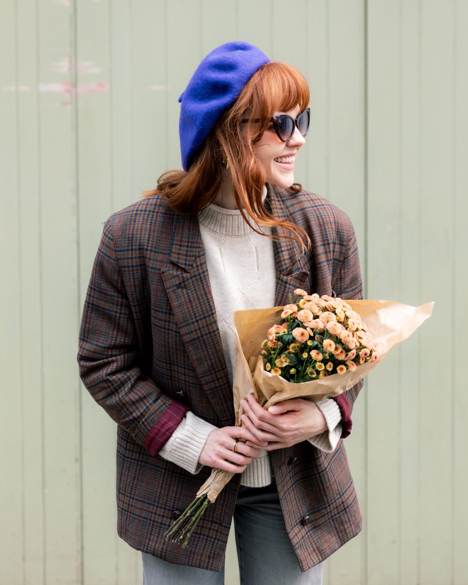 Young woman wearing a Purple Wool Beret Hat, holding a bouquet, styled with sunglasses, a cream sweater, and a plaid blazer against a paneled wall.