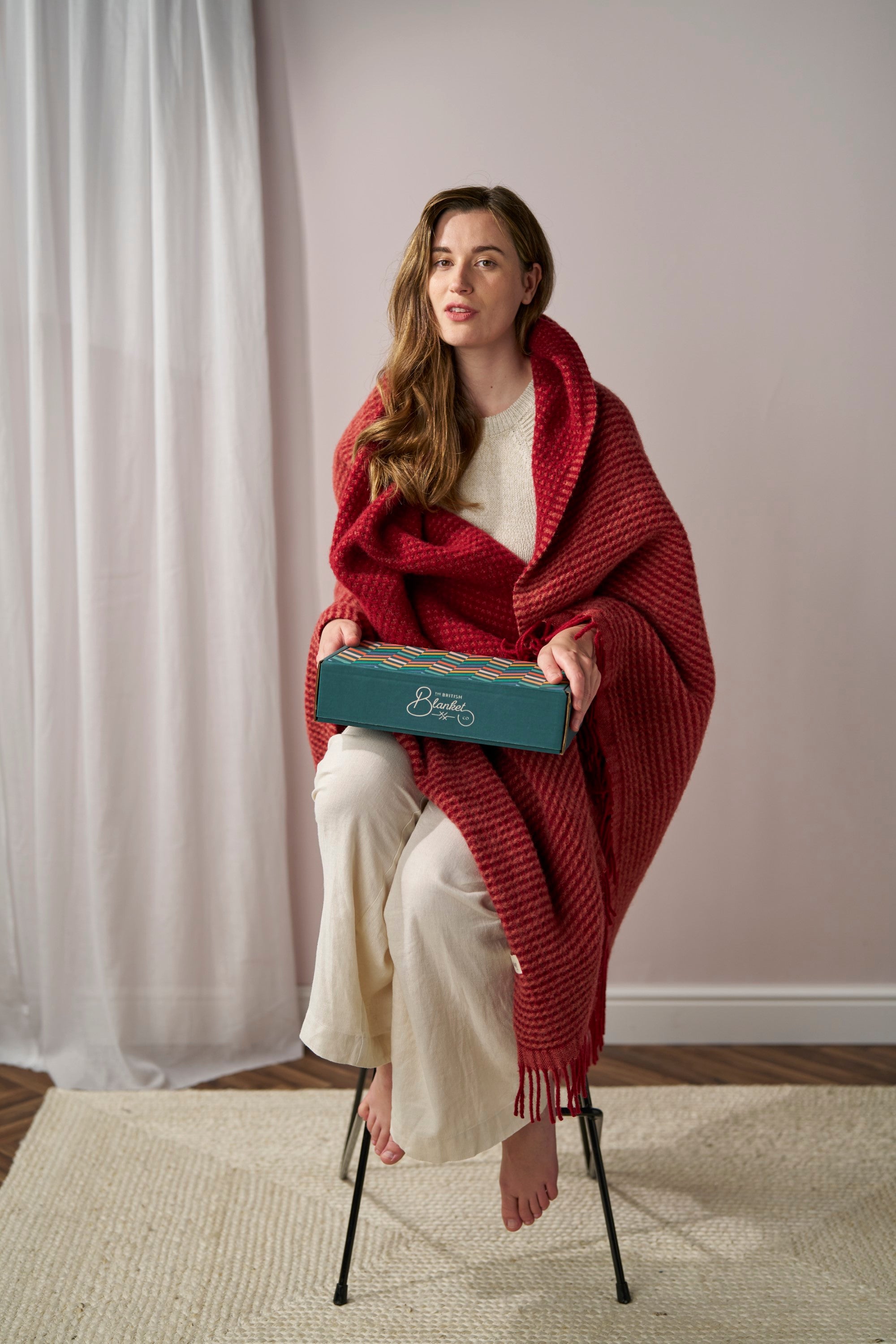 Young woman holding a forest green gift box, wrapped in a deep-red blanket. Promotes Digital Gift Card for British Blanket Company, ideal for personalized gifting.