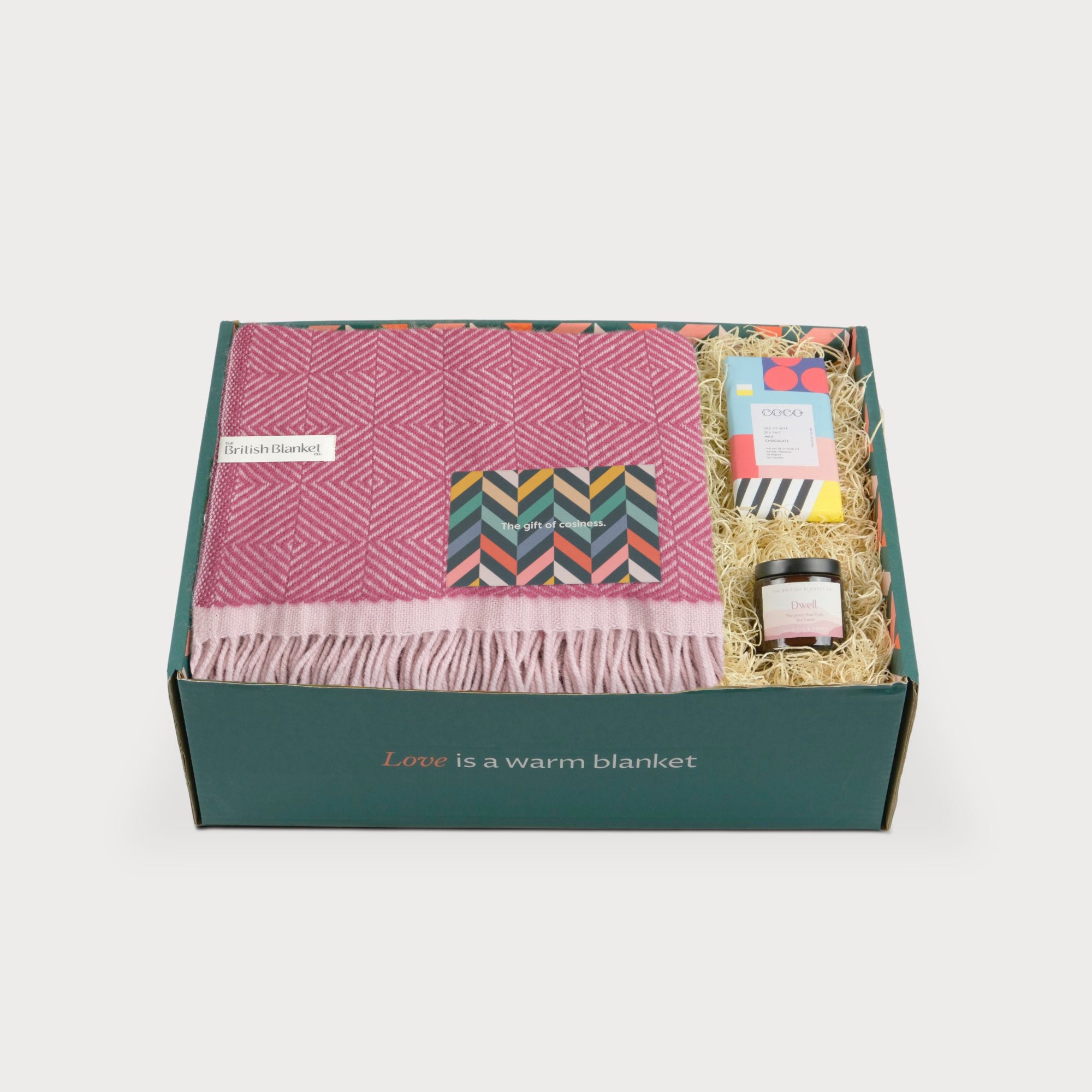Wildweave Blanket Gift Box: Clover & Briar Rose featuring a pink-and-cream throw, gourmet chocolate bar, and Dwell candle, nestled in straw with a greeting card.