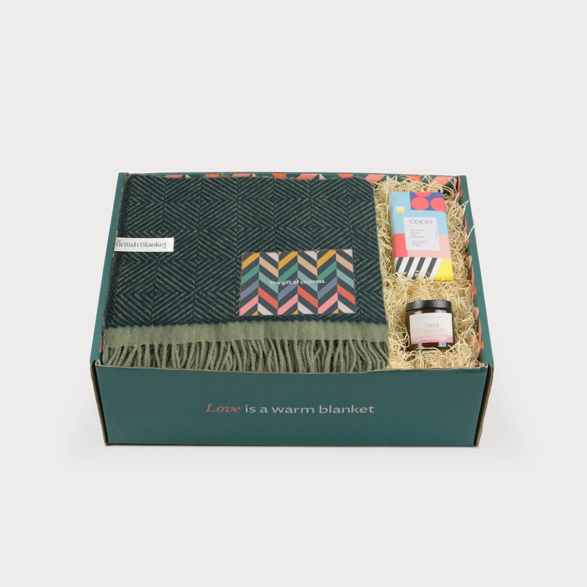 Wildweave Blanket Gift Box: Juniper & Moss includes a dark-green wool blanket, sea salt chocolate, amber jar candle, and a greeting card in a cozy arrangement.