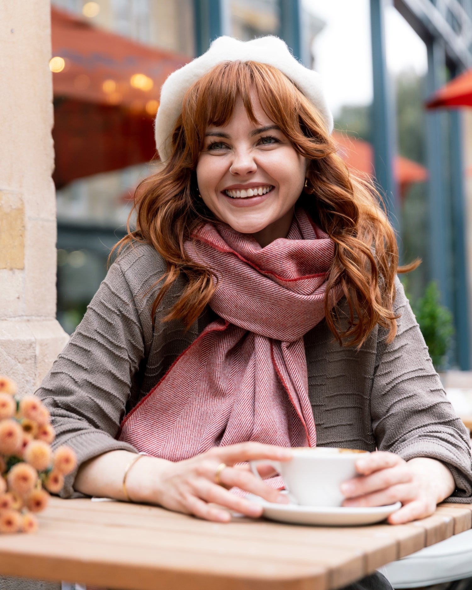 Woman in a café wearing an Oversized Blanket Scarf Gift Box: Tabasco, paired with a beret and sweater, next to a coffee cup and flowers.