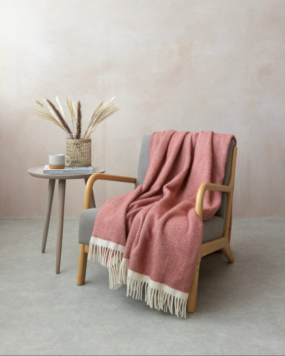 Clifton Blanket Gift Box: Rose featuring a coral-pink herringbone wool blanket draped on a chair, with a side table displaying books, a mug, and decorative grasses.