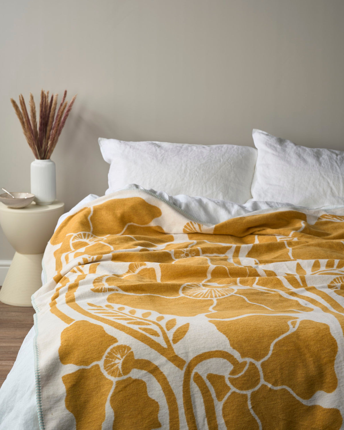 Close-up of a yellow and white floral patterned blanket on a bed with white pillows and a vase of reeds.