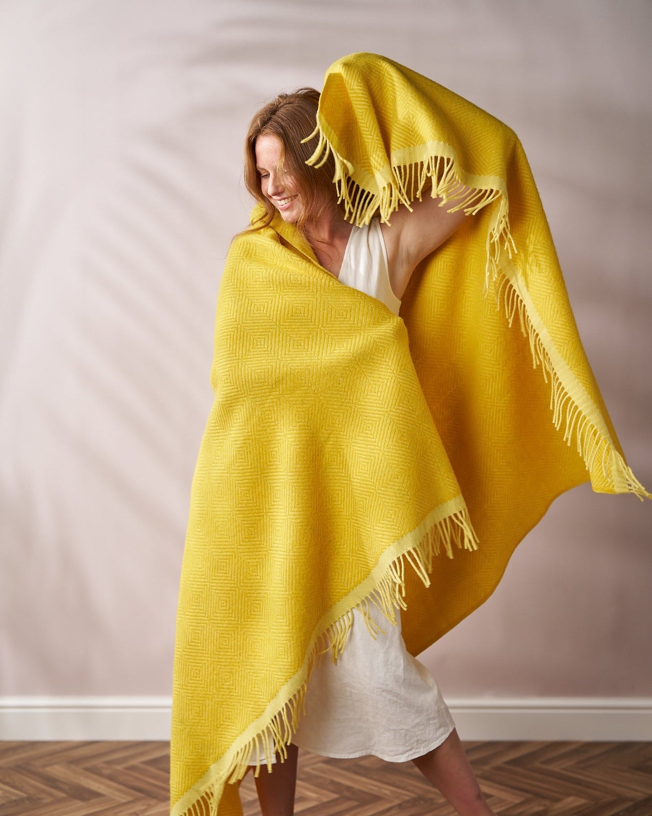 Woman joyfully drapes the Wildweave Blanket: Buttercup and Primrose, showcasing its geometric pattern and fringed edges, accentuating her light dress against a neutral backdrop.