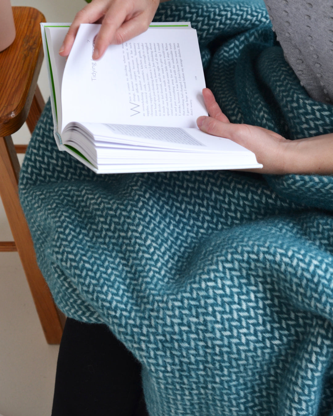 A person holding an open book. A green herringbone wool throw is draped over their lap.