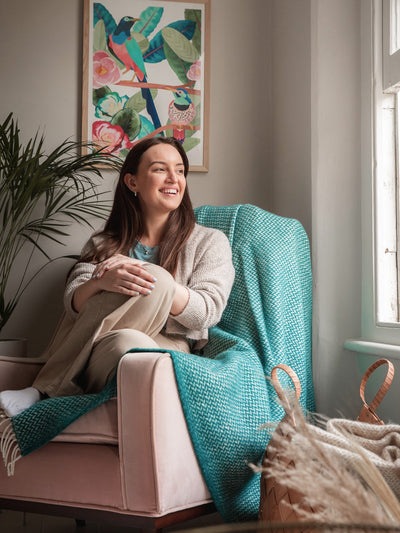 A woman sitting with her knees tucked in on a lounge chair. A green herringbone wool throw is draped over the chair.