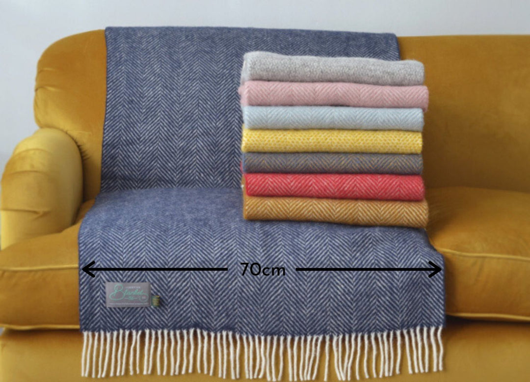 A stack of folded wool blankets on a yellow sofa. A blue blanket measuring 70 centimeters in width is draped on the sofa. 