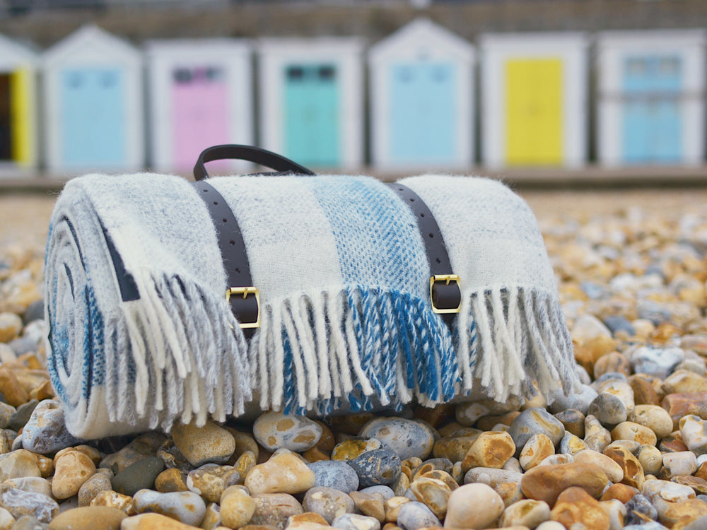 Closeup of a blue and white picnic rug on a stone beach. The picnic rug is rolled up with leather straps. 