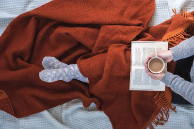A person reading a book in bed while wrapped with an extra large red wool blanket