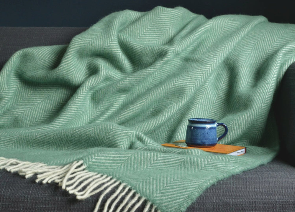Large sea green herringbone wool blanket draped over a sofa with mug and book on top the blanket