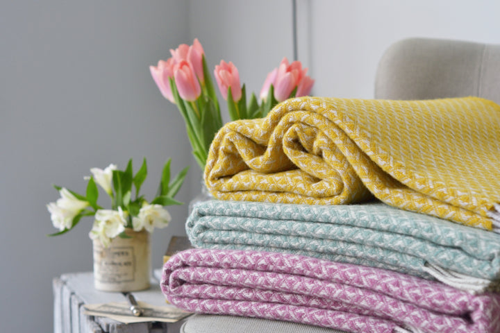 Three diamond wool blankets in yellow, blue, and pink stacked on a lounge chair