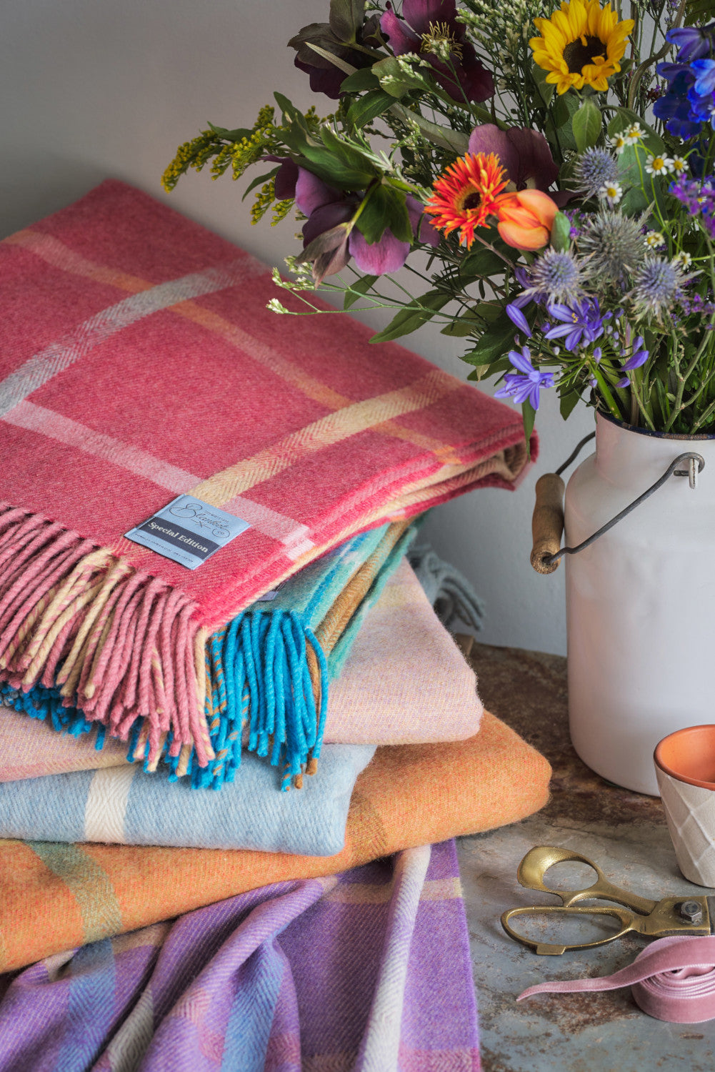 A folded red merino lambswool blanket on top a stack of wool throws beside a pot of flowers