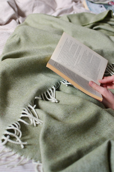A person holding an open book on top of a green merino herringbone wool blanket