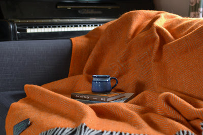 Large orange and grey herringbone wool blanket draped across a sofa with a mug and books placed on top the blanket