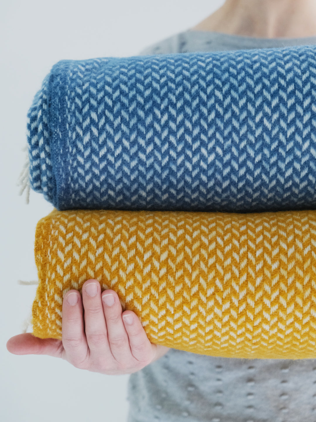 A woman holding out two herringbone wool throws in blue and yellow
