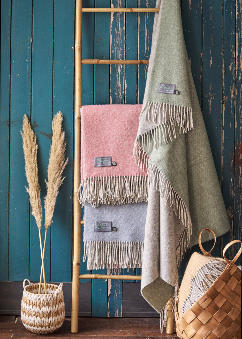 Folded green pink and blue reversible wool throw blankets by The British Blanket Company hanging on a bamboo ladder