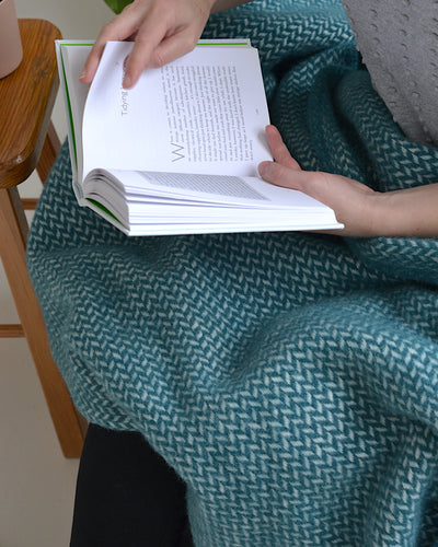 A person holding an open book. A green wool blanket is draped over their lap. 