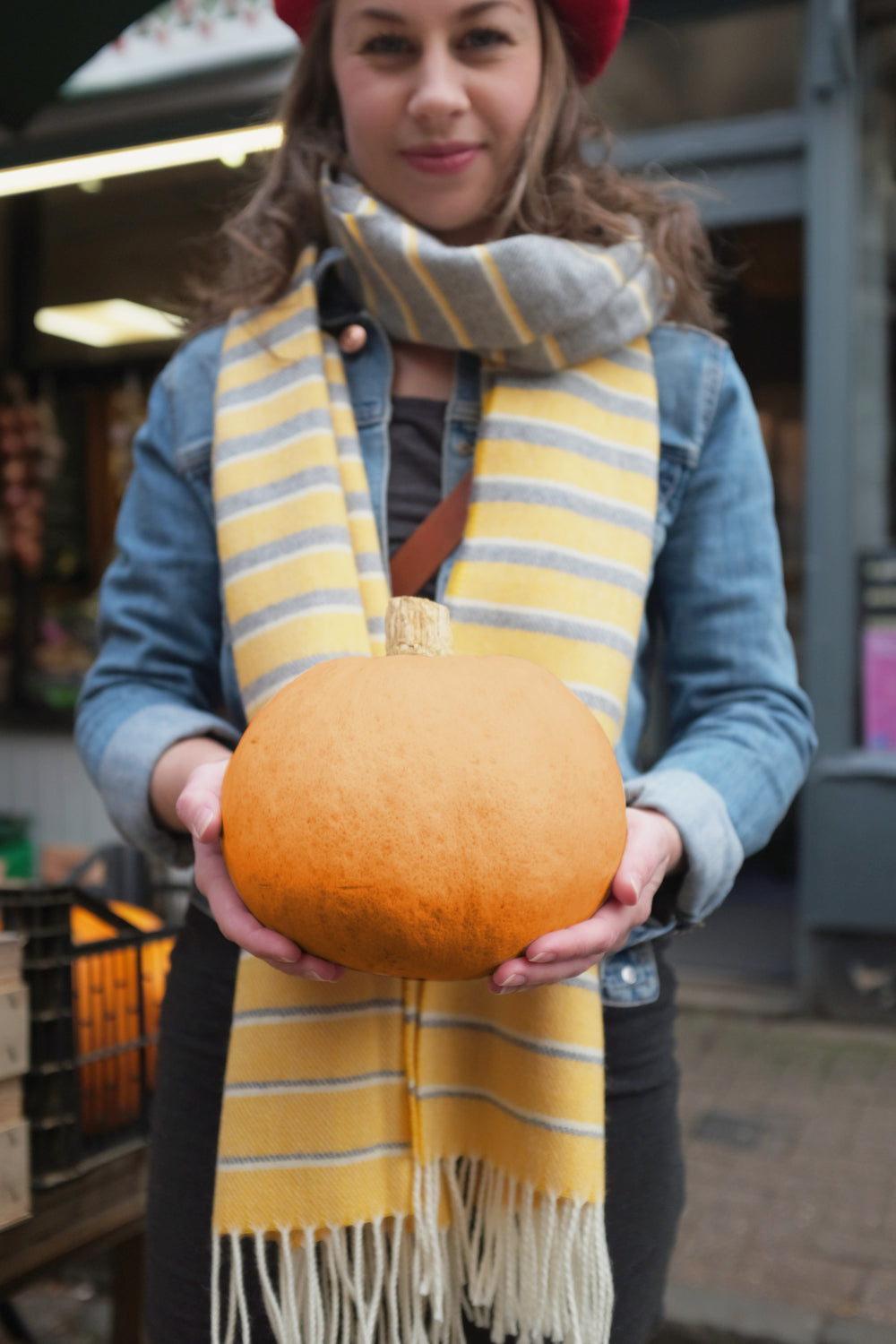 A woman wearing a yellow and grey oversized blanket scarf around her neck. The woman is holding out an orange pumpkin