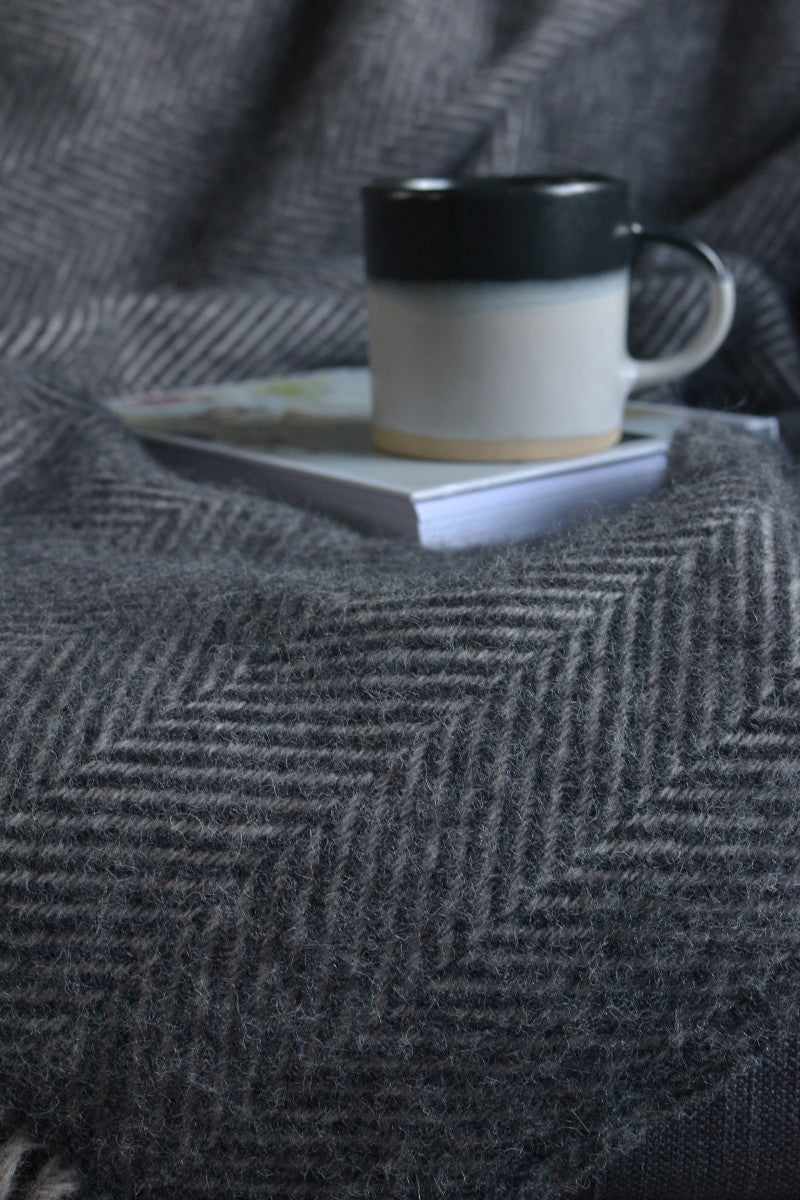 A mug and book placed on top of a grey herringbone wool blanket. 