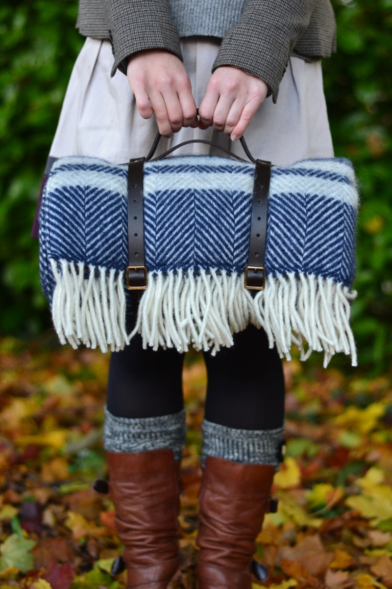 A woman holding the handle of leather straps carrying a blue and grey herringbone wool picnic blanket