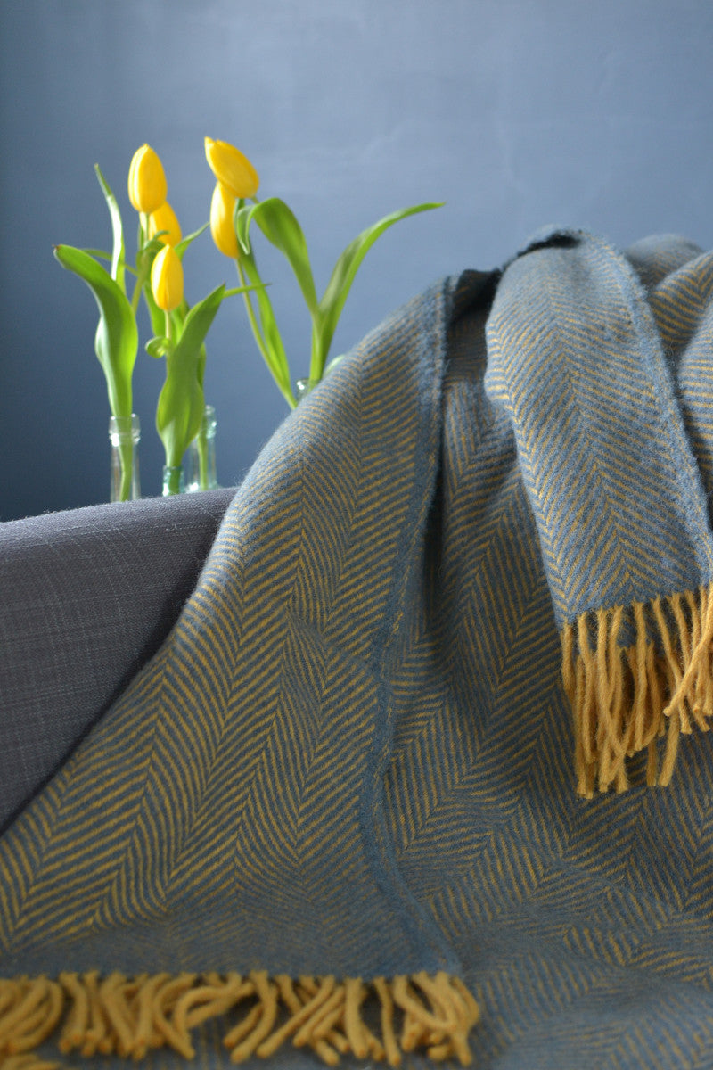 Closeup of a blue and yellow herringbone wool blanket on a sofa. Yellow flowers are placed behind the sofa