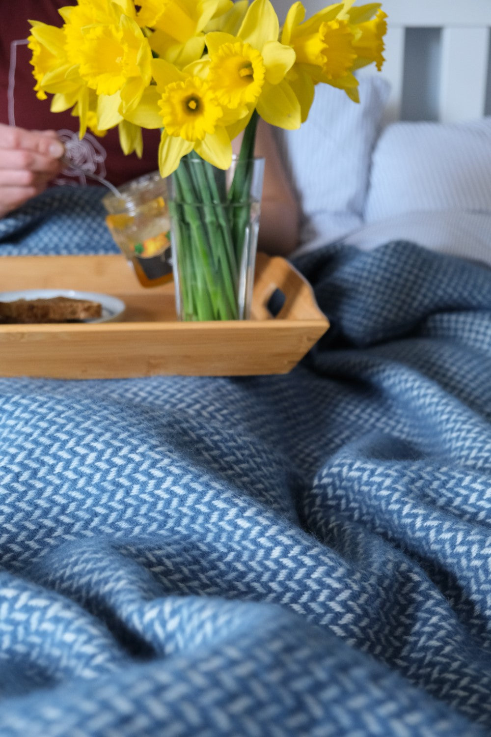 A person having breakfast in bed while underneath a blue herringbone wool blanket.