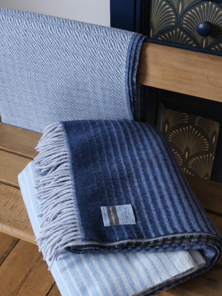 A folded blue ombre merino wool blanket on top a wooden table with a blue ripple wool throw behind it