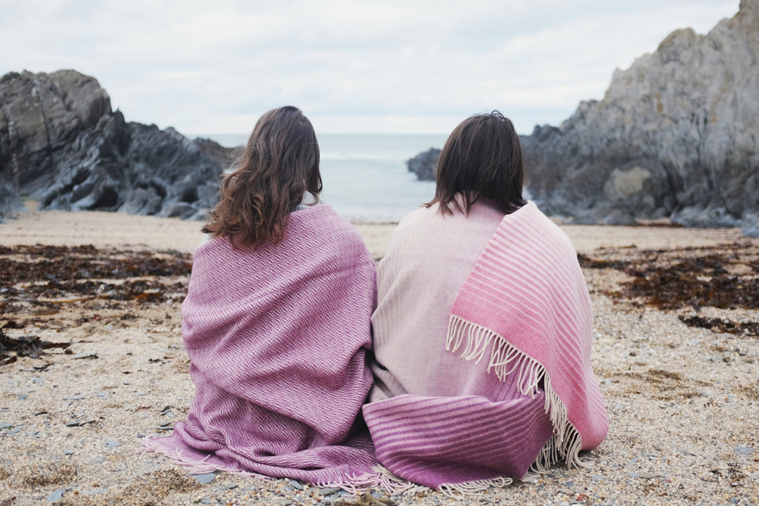 Two women sitting next to each other on the beach wrapped in pink merino wool blankets