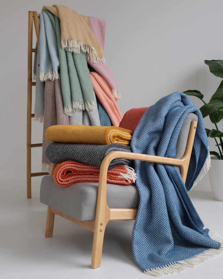 A stack of folded wool blankets on a lounge chair with a blue throw draped over the chair. Wool blankets are hanging over a wooden ladder in the background