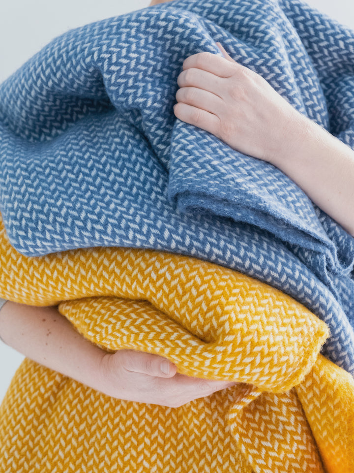 A person holding two herringbone wool blankets in blue and yellow.