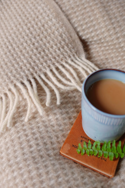 A cup of tea and a book are placed on top of an extra large cream waffle wool blanket